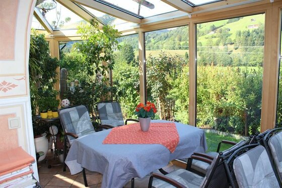 Ein heller Wintergarten mit Esstisch, Stühlen und Blick auf grüne Berghänge.