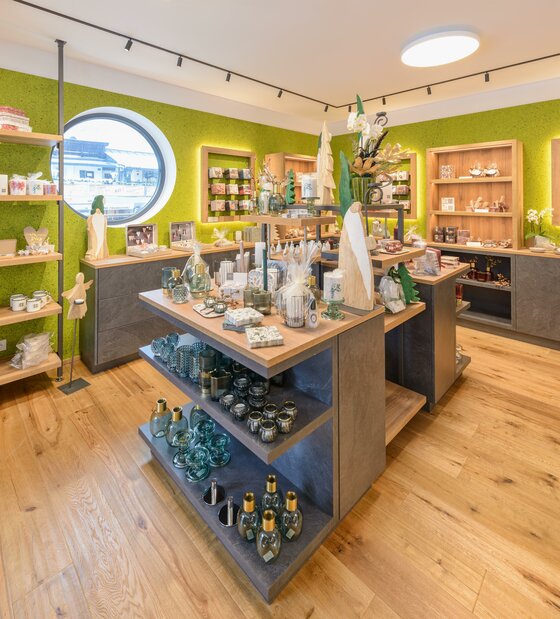 Edelwelt Kitzbühel Verkaufsraum Innenansicht eines hellen Ladengeschäfts mit Holzregalen, Glaswaren und Dekorationsartikeln vor grüner Wand.