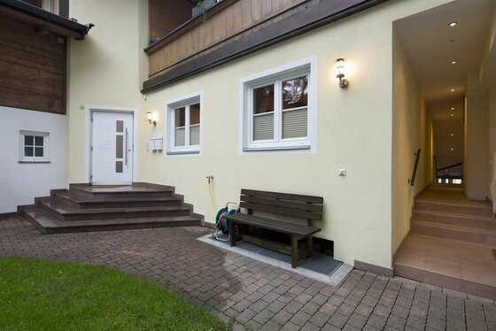 Eingangsbereich Appartment Ski Rent Michael Gepflasterter Außenbereich eines Hauses mit weißer Eingangstür, Holzbank und Treppenaufgang.