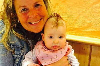 Christa Blaschke Eine lächelnde Frau hält ein Baby in den Armen. Der Hintergrund ist orangefarben, was eine freundliche Atmosphäre schafft.