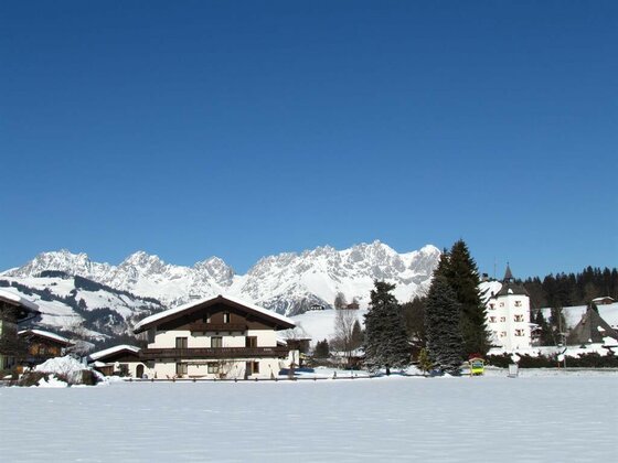 Haus Zierl im Winter Verschneites Alpenhaus vor dem Gebirgsmassiv des Wilden Kaisers unter blauem Himmel.