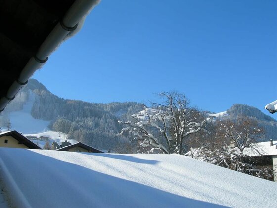 Blick über schneebedeckte Dächer auf bewaldete Berge unter blauem Himmel.