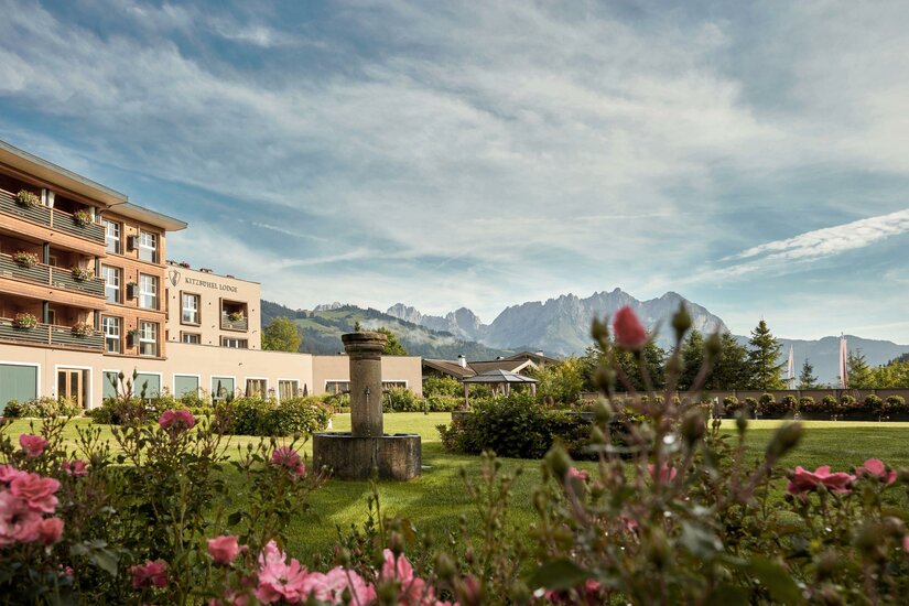 Außenansicht der Kitzbühel Lodge mit Gartenanlage, Steinbrunnen und Blick auf das Gebirge Wilder Kaiser.