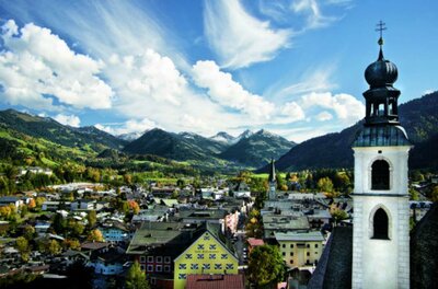 Kitzbüheler Innenstadt Sommer Eine malerische Landschaft mit einem Blick auf ein kleines Dorf umgeben von Bergen. Die Wolken am blauen Himmel verleihen der Szene eine friedliche Atmosphäre.