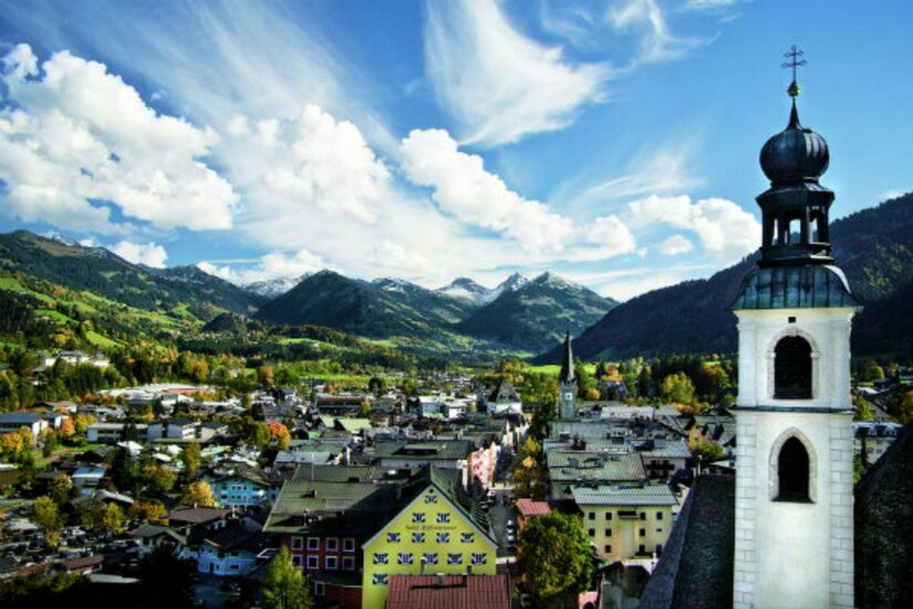 Kitzbüheler Innenstadt Sommer Eine malerische Landschaft mit einem Blick auf ein kleines Dorf umgeben von Bergen. Die Wolken am blauen Himmel verleihen der Szene eine friedliche Atmosphäre.