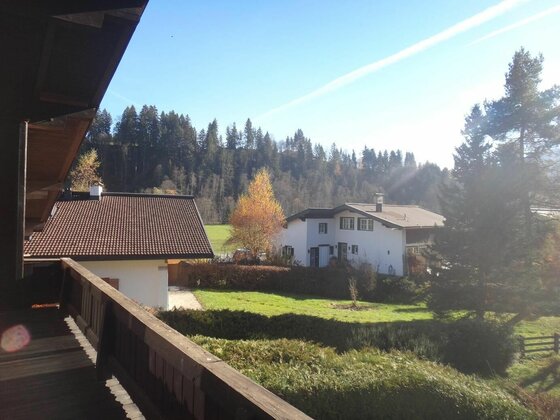 Balkonausblick Apartment Panorama Blick von einem Holzbalkon auf Wohnhäuser, eine Wiese und einen bewaldeten Hügel unter blauem Himmel.