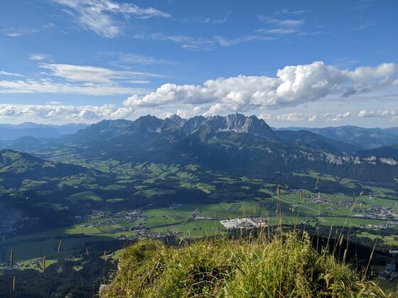 Blick von einem grasbewachsenen Gipfel auf ein weites Tal und ein schroffes Gebirgsmassiv unter blauem Himmel.