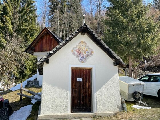 Kapelle an der Pocherrunde Kleine weiße Kapelle mit Holztür und Wandgemälde vor Nadelbäumen.