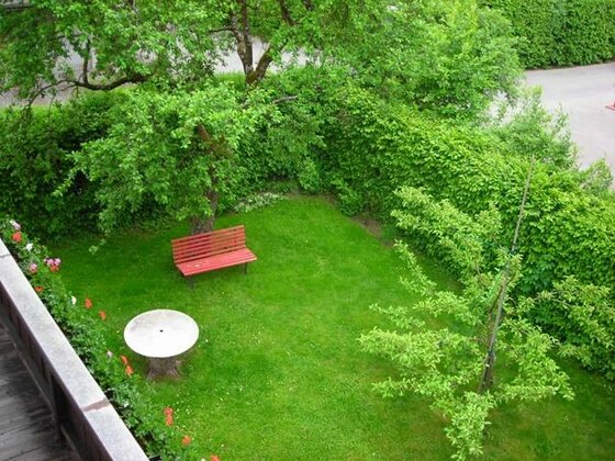 Garten des Gästehauses Maria Hilde Blick von oben auf einen grünen Garten mit roter Holzbank, rundem Steintisch und hohen Hecken.