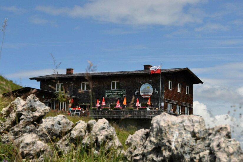 Hornköpflhütte im Sommer Zweistöckige Berghütte aus dunklem Holz mit Sonnenschirmen auf der Terrasse hinter Felsen.