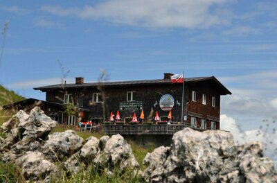 Hornköpflhütte im Sommer Zweistöckige Berghütte aus dunklem Holz mit Sonnenschirmen auf der Terrasse hinter Felsen.