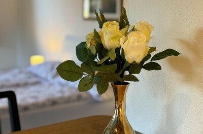 Blumendekoration im Stadl 2 Weiße Rosen in einer goldenen Vase auf einem Holztisch vor einem unscharfen Schlafbereich.
