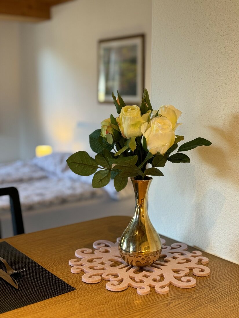Blumendekoration im Stadl 2 Weiße Rosen in einer goldenen Vase auf einem Holztisch vor einem unscharfen Schlafbereich.