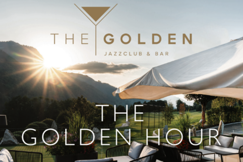 Ein eleganter Außenbereich mit Blick auf die Berge und einer sanften Sonnenuntergangsstimmung. Der Schriftzug "THE GOLDEN HOUR" kennzeichnet die entspannte Atmosphäre des Jazzclubs und der Bar.
