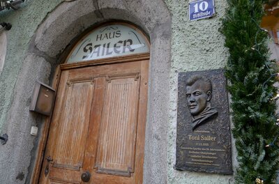 Geburtshaus Toni Sailer Eine alte Holztür mit dem Schild "Haus Sailer" und einer Gedenktafel für Toni Sailer. Die Wand ist in einem sanften Grün gestrichen.