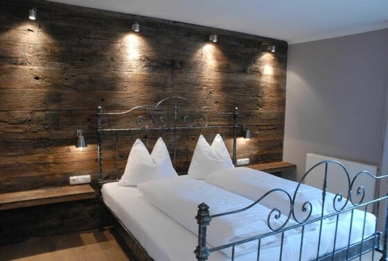 Doppelbett mit Metallgestell vor einer dunklen Altholzwand mit Spotbeleuchtung.