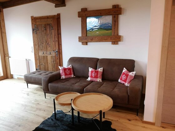 Wohnzimmer im Brandhof Braunes Sofa mit rot-weiß karierten Kissen vor einer weißen Wand mit Holzbild und Holztür.