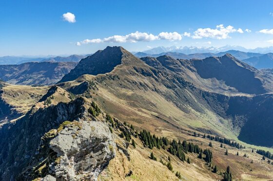 Blick auf einen grasbewachsenen Gebirgskamm mit felsigen Gipfeln unter blauem Himmel.
