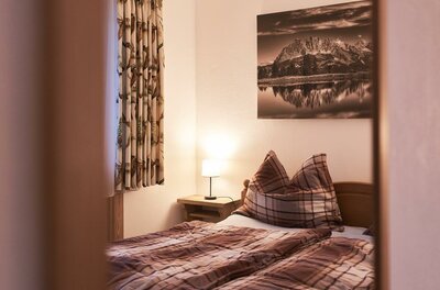 Schlafzimmer Ferienwohnung Hahnenkammblick Blick durch einen Türrahmen auf ein Doppelbett mit karierter Bettwäsche und ein Bergbild an der Wand.