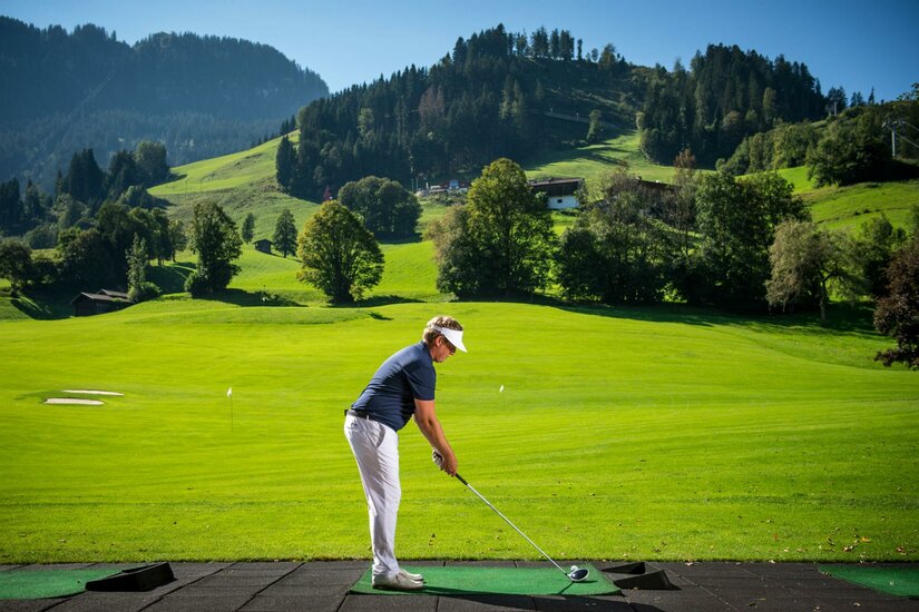 Golf Academy Ein Golfer steht auf einer Driving Range inmitten einer grünen Landschaft. Im Hintergrund sind sanfte Hügel und Bäume zu sehen.