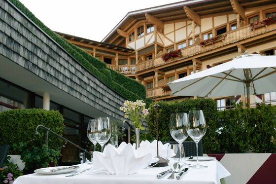 Gedeckter Tisch auf einer Hotelterrasse mit Weingläsern und Blick auf ein Holzgebäude im alpinen Stil.