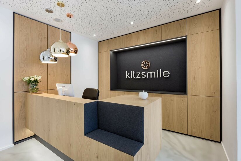 Empfangsbereich Kitzsmile Moderner Empfangstresen aus Holz mit Logo-Schriftzug an der Rückwand und drei Hängeleuchten.