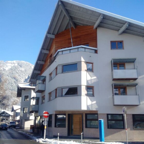 Mehrstöckiges Gebäude mit Satteldach und Balkonen vor schneebedeckten Bergen.