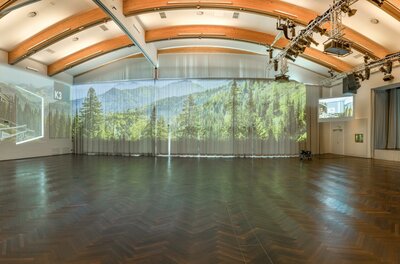 Ein moderner Veranstaltungsraum mit Holzdecke und großen Fenstern. Auf den Wänden sind Landschaftsprognosen und Präsentationen projiziert.
