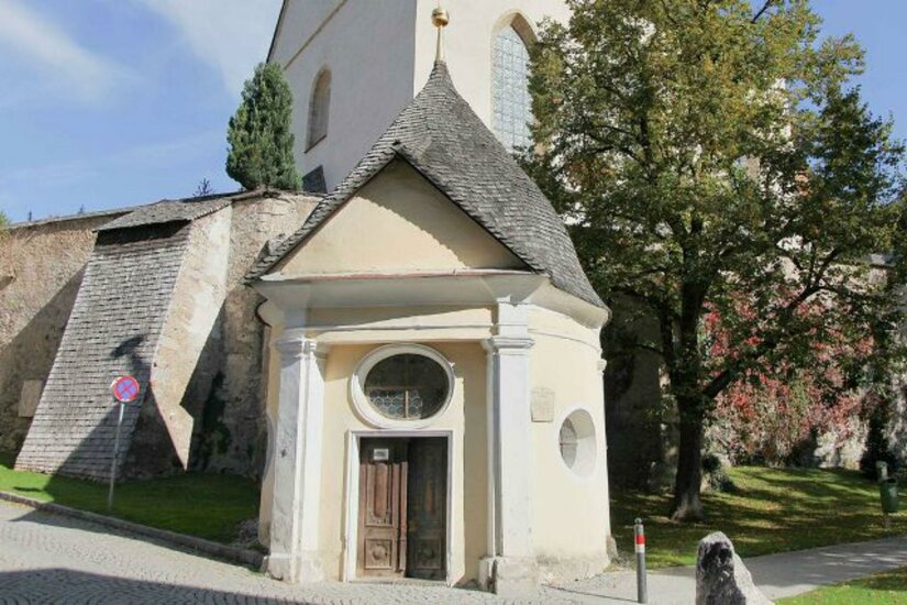 Ein kleines, historisches Gebäude mit einem spitzen Dach steht neben einer Kirche. Die Umgebung ist grün, mit Bäumen und einem ruhigen Weg.