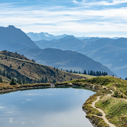 Ein klarer Bergsee spiegelt die umliegenden Berge und Wälder wider, während die sanften Hügel im Herbstlicht erstrahlen.
