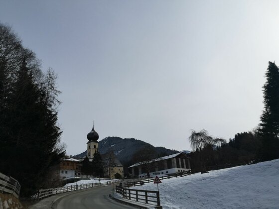 Ortsansicht von Buchau Blick auf eine asphaltierte Straße, eine Kirche mit Zwiebelturm und schneebedeckte Hänge vor bewaldeten Bergen.