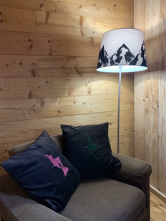 Sessel mit zwei dunklen Kissen vor einer Holzwand neben einer Stehlampe mit Bergmotiv.