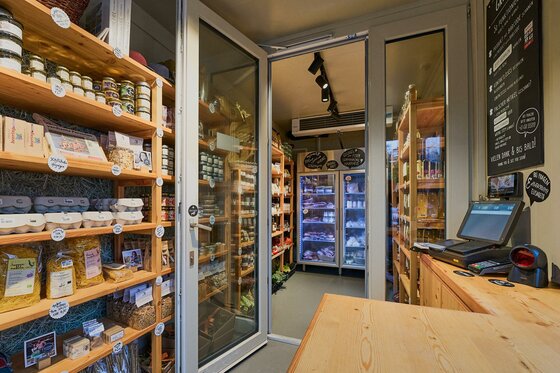 Hofladen Unsere Kiste Innenansicht eines kleinen Hofladens mit Holzregalen, regionalen Produkten und einem Selbstbedienungsterminal.