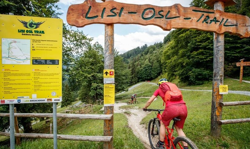 Einstieg Lisi Osl Trail Mountainbiker fährt unter einem Holzportal mit der Aufschrift Lisi-Osl-Trail in einen schmalen Pfad ein.