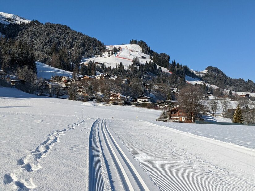 Stadtloipe Kitzbühel-Aurach-Jochberg Präparierte Langlaufloipe im Schnee vor einer Siedlung und bewaldeten Bergen unter blauem Himmel.