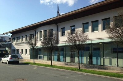 Zweistöckiges Gesundheitszentrum mit Glasfront im Erdgeschoss, davor geparktes Auto und junge Bäume.