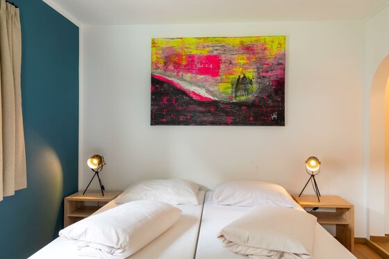 Schlafzimmer im Safestay Kitzbühel Doppelbett mit weißer Bettwäsche, Nachttischen und einem abstrakten Gemälde an der Wand.