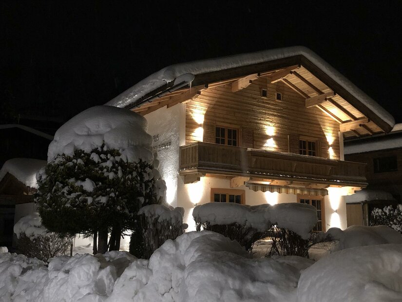 Beleuchtetes Holzhaus bei Nacht mit schneebedecktem Dach und tief verschneiten Hecken im Vordergrund.