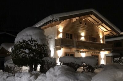 Haus Hagleitner im Winter Beleuchtetes Holzhaus bei Nacht mit schneebedecktem Dach und tief verschneiten Hecken im Vordergrund.