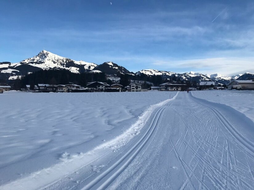Sonnenloipe Golfplatz Runde Präparierte Langlaufloipe auf einer verschneiten Ebene vor einer Bergkulisse und Häusern.