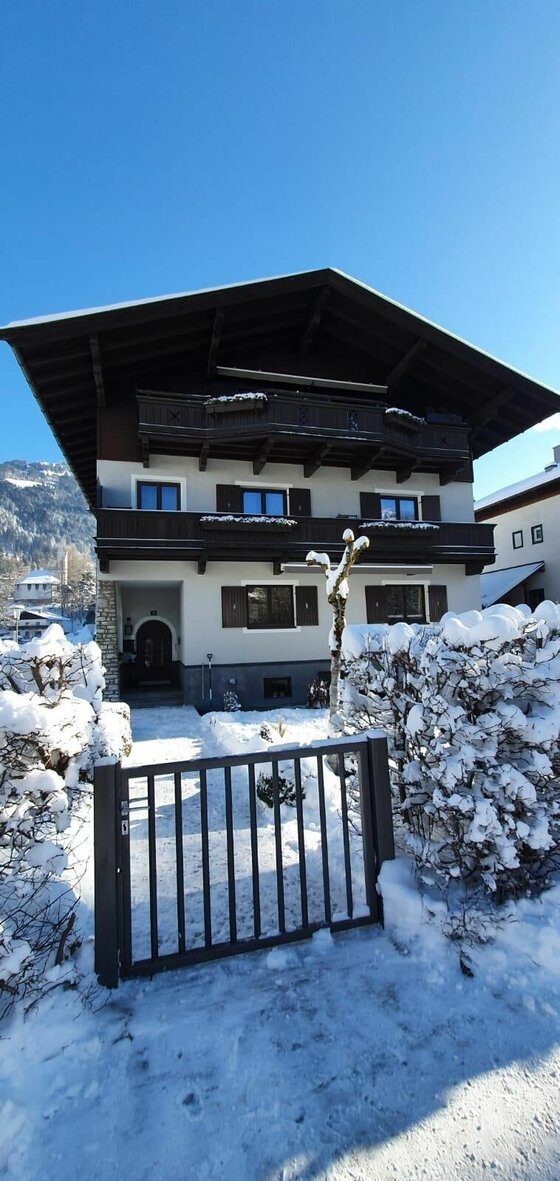 Mehrstöckiges alpines Wohnhaus mit Holzbalkonen im Winter vor blauem Himmel.