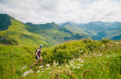 Eine Frau läuft einen grasbewachsenen Bergpfad hinauf, im Hintergrund eine weite Gebirgslandschaft unter bewölktem Himmel.