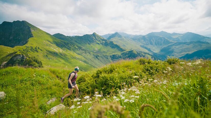 Eine Frau läuft einen grasbewachsenen Bergpfad hinauf, im Hintergrund eine weite Gebirgslandschaft unter bewölktem Himmel.