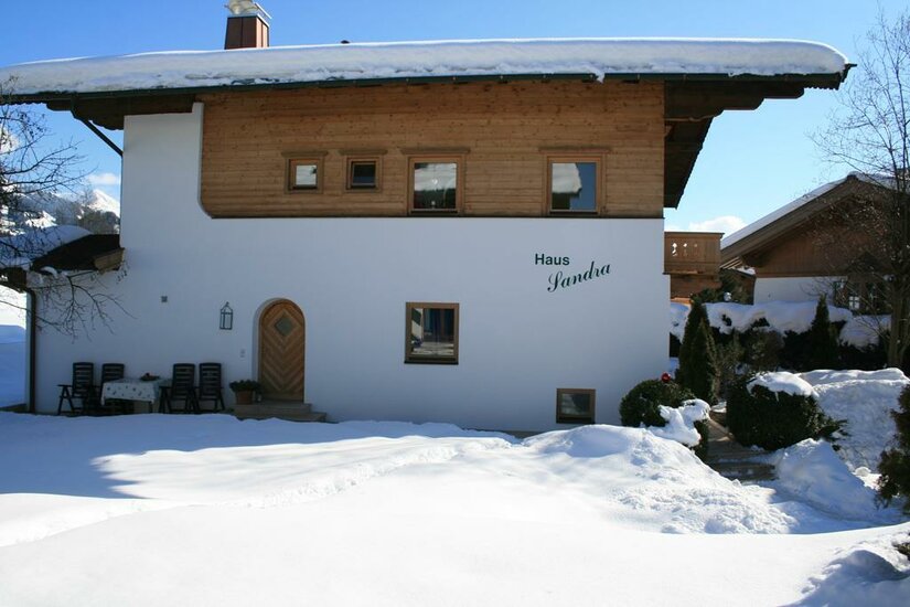 Haus Sandra im Winter Zweistöckiges Haus mit weißer Fassade, Holzobergeschoss und schneebedecktem Dach in einer Winterlandschaft.