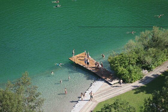 Ein klarer grüner See mit Menschen, die schwimmen und auf einem Holzsteg stehen. Umgeben von Bäumen und einer schönen Landschaft.