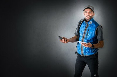 Ein Mann in einer blauen Weste steht vor einem grauen Hintergrund. Er hält ein mobiles Gerät in einer Hand und ein Tablet in der anderen.