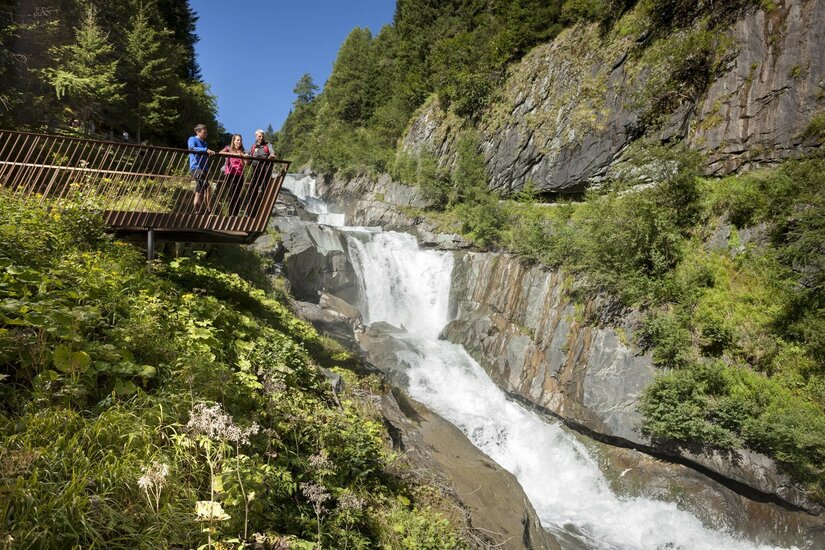 Das Bild zeigt die Umbalfälle mit einem kraftvollen Wasserfall, der durch eine felsige Schlucht fließt. Links befindet sich eine Aussichtsplattform aus Metall, auf der mehrere Personen stehen und den Wasserfall betrachten. Die Schlucht wird von steilen Felswänden, dichtem Grün und hohen Bäumen begleitet.