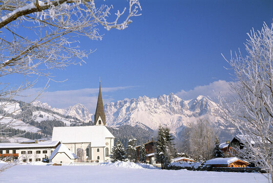 Verschneite Kirche vor einem Gebirgsmassiv unter blauem Himmel mit frostigen Zweigen im Vordergrund.