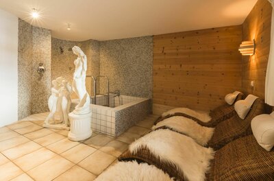 Wellnessbereich mit Ruheliegen, Fellauflagen, einem kleinen Tauchbecken und weißen Statuen vor einer Holzwand.