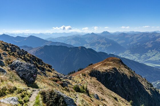 Blick von einem felsigen Bergpfad auf eine weite Gebirgslandschaft unter blauem Himmel.